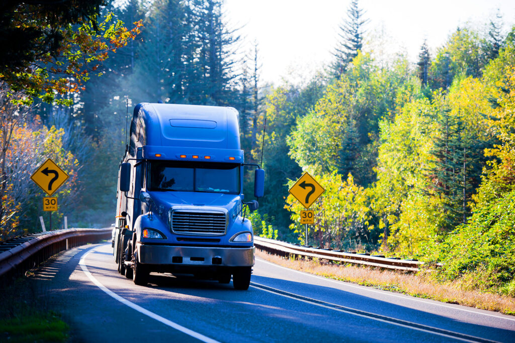 Blue semi truck driving on winding highway through autumn forested road