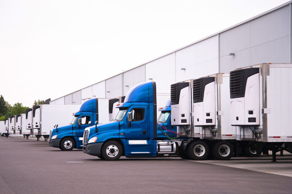 Fleet of blue semi-trucks with refrigerated trailers parked at commercial warehouse loading dock